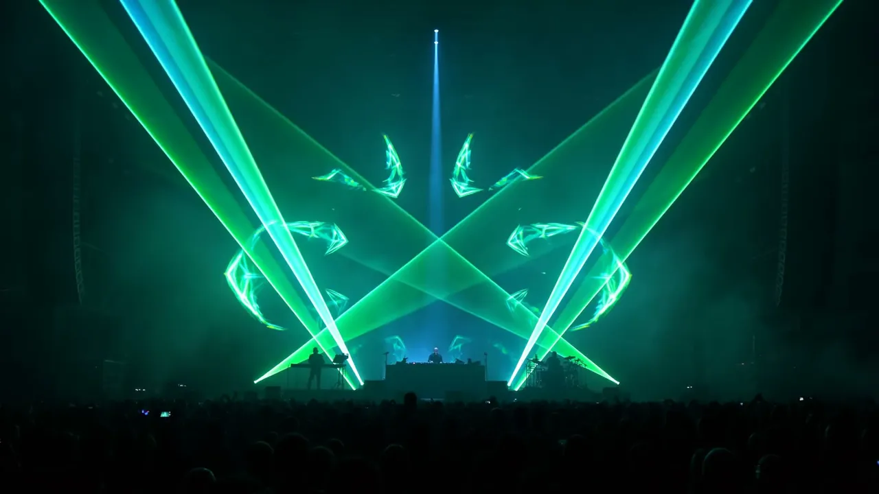 Two DJs on a stage under crossing green laser fans, with hands up in the crowd below.