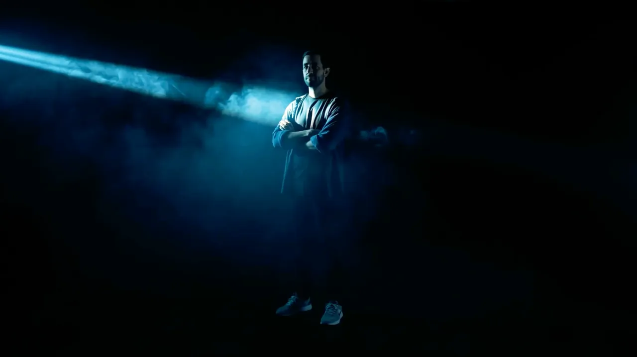 A person standing in a dark studio, lit by a single blue beam through haze.